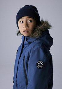 Blå hættetrøje med pelskant, med sorte lynlåslommer og et rundt logo-patch på venstre ærme. Båret med en mørk hue.