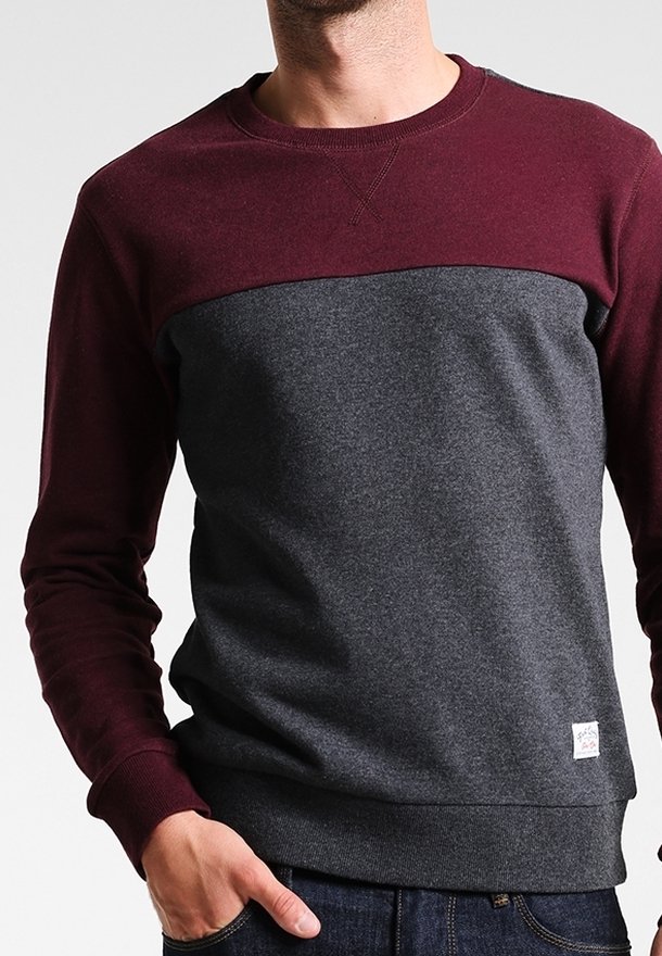 Homme portant un sweat-shirt à manches longues et col rond avec une partie supérieure bordeaux et une partie inférieure gris foncé, main dans la poche du jean.