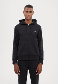 Black zip-up hoodie with a hood, featuring white logo, side stripes, and ribbed cuffs. Made of smooth, elastic fabric, suitable for casual wear.