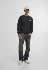Zwarte sweatshirt met geribde manchetten, lichtbeige pet en donkerblauwe jeans. Sneakers met beige accenten. Eenvoudige, casual outfit.