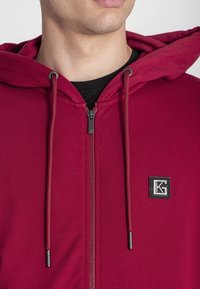 Crvena zip-up majica s kapuljačom napravljena od mekog materijala, s kapuljačom s utezima, prednjim patentnim zatvaračem i malim crnim logotipom na prsima.