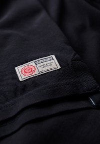 Tela negra con una superficie texturizada que presenta una etiqueta tejida rectangular. La etiqueta incluye "SUPERDRY" y "VINTAGE ATHLETIC OUTFITTERS" en rojo y azul.
