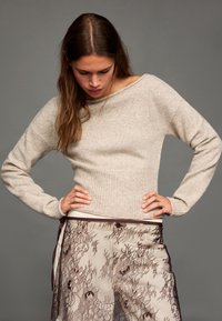 Világosbézs kötött pulóver hajónyakrésszel és bordázott szegéllyel. Áttetsző csipkenadrággal párosítva, amely virágmintákkal és kontrasztos részletekkel díszített.