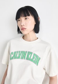 Cremefarvet bomulds t-shirt med korte ærmer, der har et grønt "CALVIN KLEIN" logo i en buet, fed skrifttype. Enkel og uudsmykket design.
