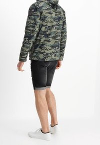 Giacca con motivo camouflage in tessuto verde e nero, design con cappuccio, abbinata a pantaloni denim neri aderenti e sneakers bianche.