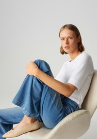 T-shirt blanc côtelé, jean bleu à jambes larges et ballerines beiges. Modèle assis sur une chaise de couleur claire avec un fond neutre.