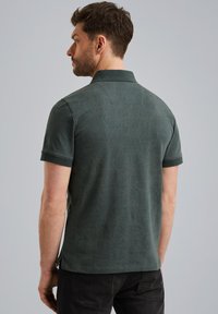 Mørkegrøn polo-shirt med et struktureret mønster, korte ærmer og en klassisk krave. Sideslidser ved kanten tilføjer detalje. Bæres med mørke jeans.