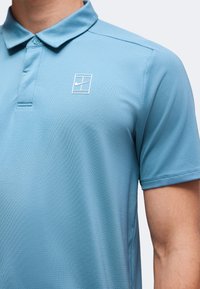 Homme portant un polo bleu clair à manches courtes avec un logo Nike blanc dans un carré sur le côté gauche de la poitrine.
