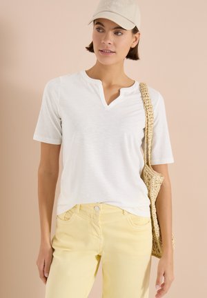 Femme portant un haut blanc à manches courtes, un pantalon jaune, une casquette beige avec le texte « OPTIMISME » et portant un sac à épaule tressé, sur un fond beige.