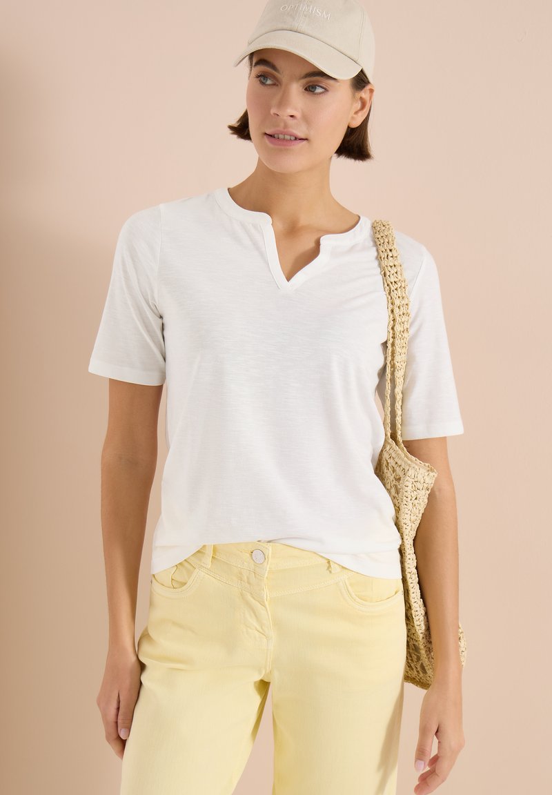 Femme portant un haut blanc à manches courtes, un pantalon jaune, une casquette beige avec le texte « OPTIMISME » et portant un sac à épaule tressé, sur un fond beige.