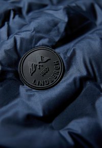 Marinblått tyg med veckad textur har en cirkulär svart logotyp präglad med "LINDENBERGH" och "1927", som föreställer berg- och flaggmotiv.