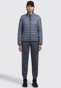 Polstret grå jakke med bølgemønster, høj krave og lynlås foran, parret med mørkegrå sweatpants og sorte sneaks.