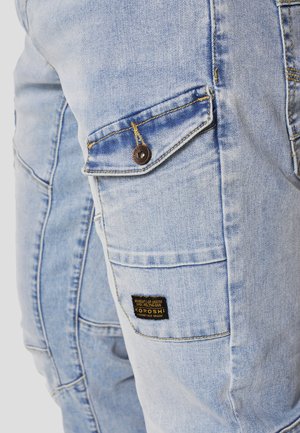 Hele sinised teksapüksid, millel on nööpidega klapiga tasku ja must silt, millel on kirjutatud "Koroshi", reie külje peal.