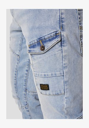 Hele sinised teksapüksid, millel on nööpidega klapiga tasku ja must silt, millel on kirjutatud "Koroshi", reie külje peal.