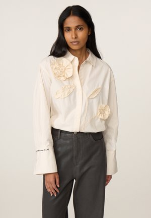 Vrouw draagt een crèmekleurig overhemd met knopen en stoffen bloem- en bladdetails, gecombineerd met hooggetailleerde zwarte broek, staand tegen een effen achtergrond.
