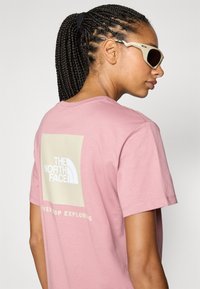 Rosa kortärmad t-shirt med en stor vit logotyp och en beige rektangulär bakgrund. Slät tygkvalitet, avslappnad passform, vardaglig design.