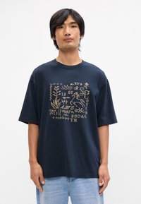 Μπλέ σκούρο βαμβακερό μπλουζάκι με χρυσό γραφικό σχέδιο που περιλαμβάνει μοτίβα φύσης και κείμενο. Χαλαρή εφαρμογή με κοντά μανίκια και στρογγυλή λαιμόκοψη.