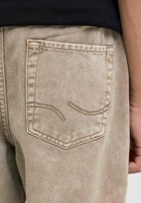 Bež hlače z zadnjim žepom in subtilnim ukrivljenim šivom, narejene iz teksturirane tkanine, s poudarkom na vzdržljivosti in sproščenem slogu.