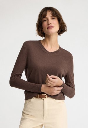 Kvinde med kort brunt hår iført en brun langærmet V-hals trøje, beige bukser og et brunt bælte, poserer med hænderne foldet.