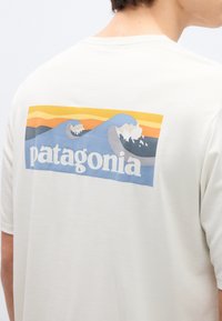 Άτομο που φοράει λευκό μπλουζάκι με πολύχρωμο ορθογώνιο λογότυπο που απεικονίζει στυλιζαρισμένα κύματα του ωκεανού και ηλιοβασίλεμα στην πλάτη.