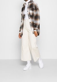 Osoba oblečená v hnědo-bílém kostkovaném tričku přes bílý rolák, smetanových širokých kalhotách, bílých ponožkách a bílých teniskách na jednobarevném pozadí.