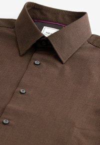 Chemise marron texturée à boutons avec un col pointu et une étiquette de marque visible à l'intérieur de l'encolure.