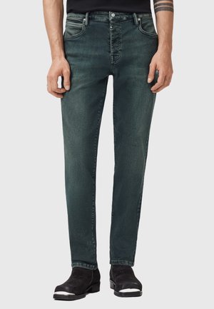 Mann trägt verblasste dunkelgrüne Slim-Fit-Jeans und schwarze Knöchelhohe Stiefel mit silbernen Spitzen, steht vor einem schlichten hellen Hintergrund.