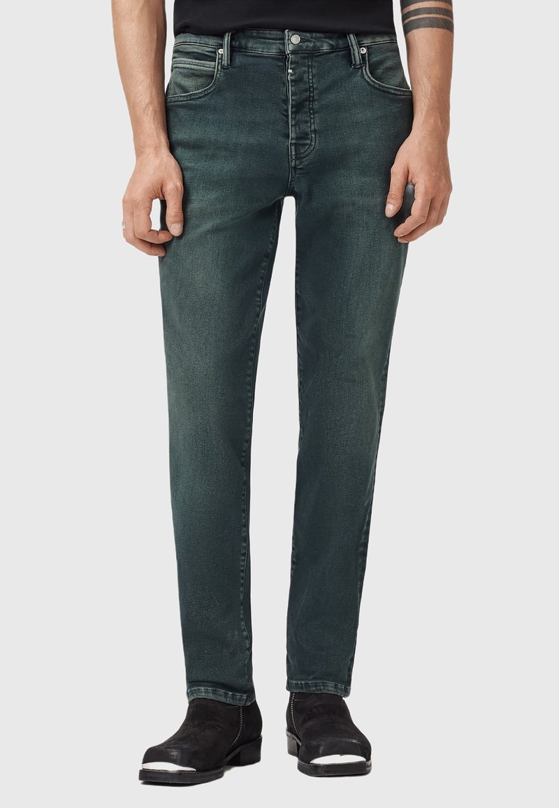 Muž v zelených úzkých džínách se zesvětleným efektem a černých kotníkových botách se stříbrnými špičkami, stojící proti jednoduchému světlému pozadí.