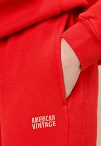 Sarkans kokvilnas treniņbikses ar iešūtu kabatu un "American Vintage" logo gaismi pelēkā krāsā, ar mīkstu, teksturētu auduma apdari.