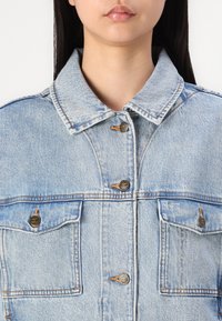 Chaqueta de denim de color azul claro con cuello, que cuenta con dos bolsillos frontales, botones de bronce y detalles de costura visibles.