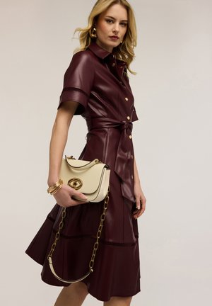 Donna che indossa un vestito di pelle bordeaux con bottoni e cintura in vita, tiene una borsa crema con catena dorata e indossa braccialetti e orecchini a cerchio dorati.