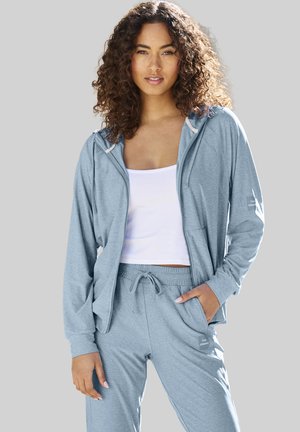 Woman with curly hair wearing a light gray zip-up hoodie, matching drawstring pants, and a white tank top, standing against a gray background.