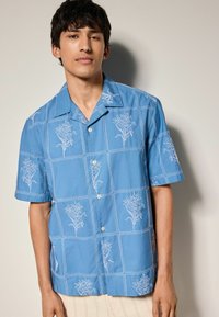 Blauw kortemouw shirt met witte bloemenborduring in een ruitpatroon, met een knoopsluiting aan de voorkant en een zachte, gladde textuur.