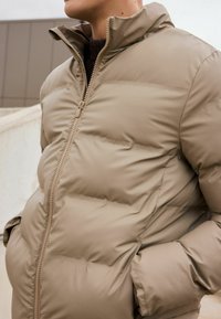 Jachetă puffer bej cu guler înalt, fermoar frontal și buzunare laterale. Textură netedă, captusită, cu detalii cusute de-a lungul cusăturilor.