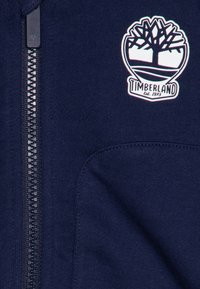 Felpa zip-up blu navy con tessuto testurizzato, caratterizzata da un logo Timberland bianco e una grafica ad albero sul lato sinistro del petto. La zip si estende lungo la parte anteriore.