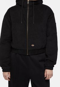 Black corduroy zip-up jacket with hood, elastic cuffs, chest pocket on sleeve, and a small logo patch above the waistband.