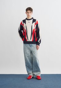 Giovane in piedi che indossa un maglione a motivi rossi, bianchi e blu navy, jeans larghi blu sbiaditi e sneakers rosse su un tappeto blu.