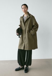 Olivgrüne Kapuzenjacke mit Kordelzug, weiter Schnitt, große Taschen, kombiniert mit schwarzen Hosen und schwarzen Loafers auf hellem Boden.