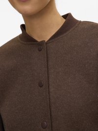 Gros plan sur une personne portant une veste en laine brune avec un col côtelé et trois boutons-pression visibles.