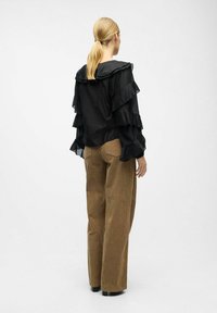 Femme avec une queue de cheval blonde portant un blouse noire à volants à manches longues et un pantalon en velours côtelé marron à jambes larges, se tenant devant un fond uni.