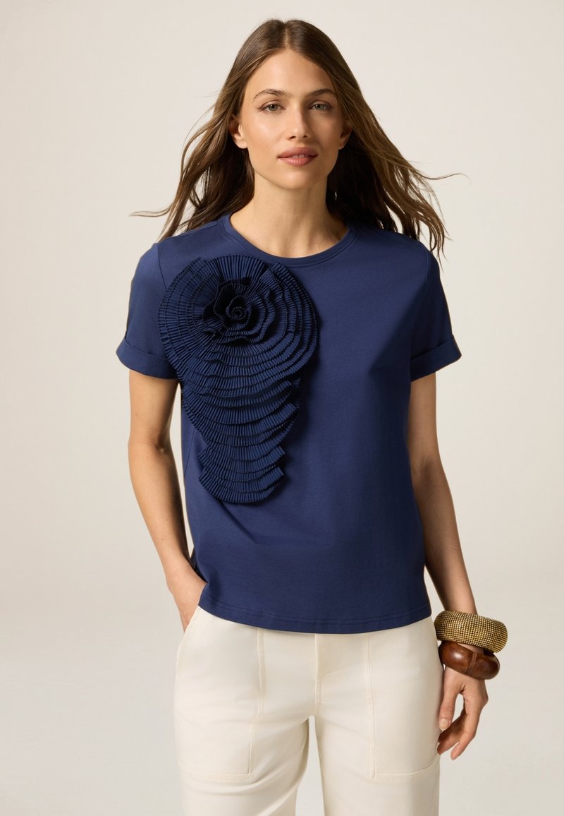 Navyblaue Baumwoll-T-Shirt mit einem strukturierten, geschichteten Stoffblumenakzent an der linken Schulter. Kurze Ärmel und ein rundes Ausschnitt.