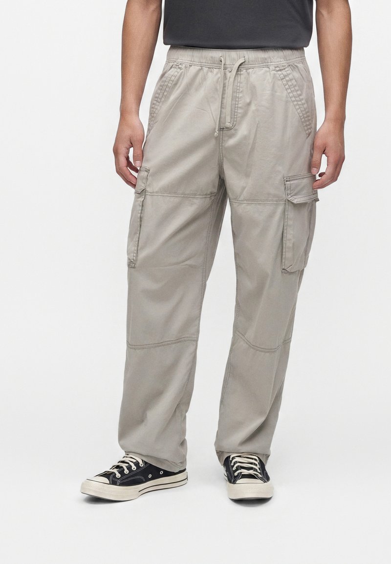 Homme portant un pantalon cargo gris clair avec des poches avant et latérales, associé à des baskets en toile noires et blanches, debout sur fond blanc.