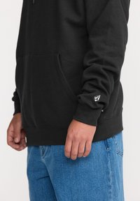 Haut à capuche noir avec une poche avant, des poignets côtelés et un petit logo sur la manche. Porté avec un jean en denim bleu, affichant une coupe décontractée.