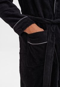 Robe nera con texture, dotata di una cintura, presenta una tasca con piping grigio e maniche dettagliate. Il tessuto sembra morbido e assorbente.