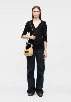 Vrouw draagt een zwart topje met V-hals en zwarte broek met wijde pijpen, houdt een beige geweven handtas met een zwarte band over haar schouder, staat tegen een witte achtergrond.