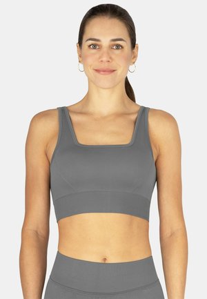 Frau mit zurückgebundenem Haar, die Creolen trägt, grauer Sport-BH mit quadratischem Ausschnitt und passende hoch taillierte Leggings, schaut nach vorne und lächelt.