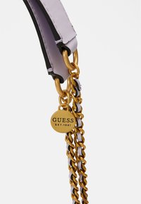 Gros plan sur une bandoulière chaîne en or tissée de cuir lavande avec un pendentif rond en or gravé de "GUESS EST. 1981."