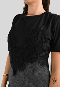 T-shirt nero sovrapposto a un top in pizzo nero. Il top presenta un intricato design in pizzo. Tessuto a quadri grigi nella parte inferiore.