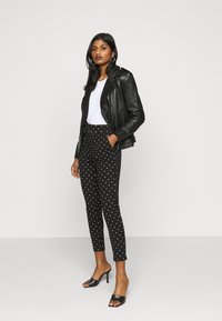 Jaqueta de couro preta, t-shirt branca, calças pretas com bolinhas e mules pretas de salto. O visual apresenta um corte slim, tecido texturizado e um estilo casual.
