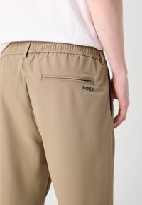 Khaki sporta bikses ar elastīgu jostasvietu, sānu kabatu un rāvējslēdzēja aizmugurējo kabatu ar melnu logotipa akcentu. Gluda tekstūra.